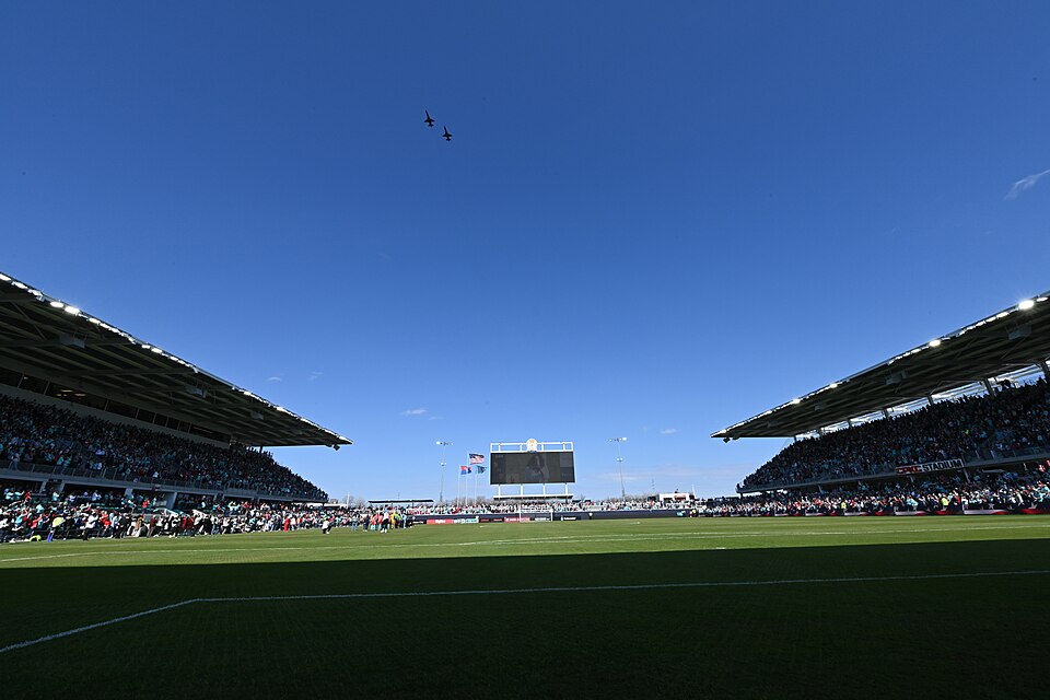 Two fighter jets fly over a packed soccer stadium with a large scoreboard.