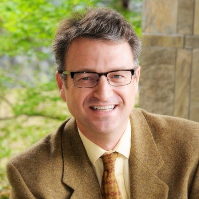 Speaker at a conference event, wearing glasses and a brown jacket.